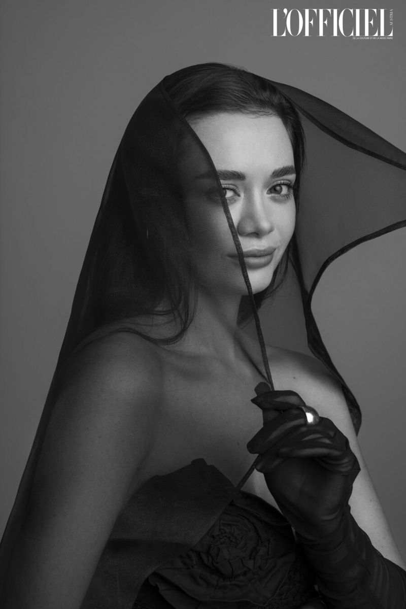 A portrait of a brunette woman wearing a black veil and black gloves, set against a neutral grey backdrop in a studio setting. Fashion.
