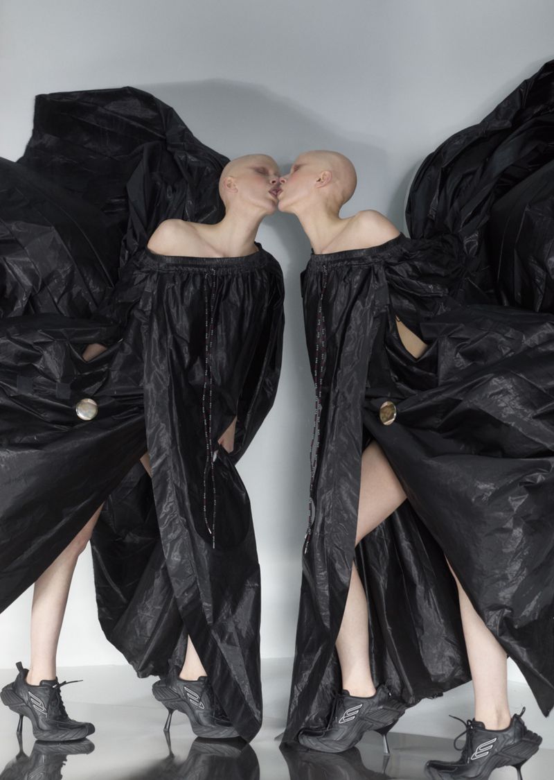 Two bald adult women in oversized black gowns kiss in a light studio. The fashion ensemble includes platform heeled shoes. 