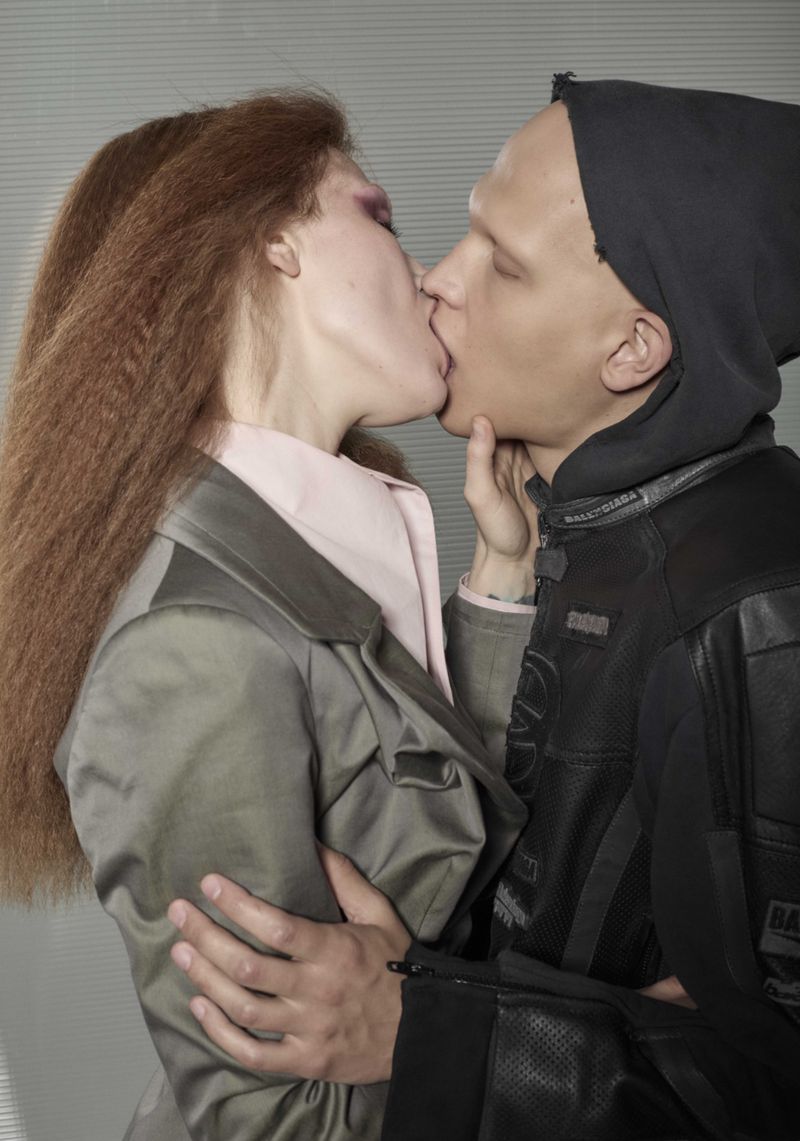Close-up studio portrait of a couple kissing; the female has auburn crimped hair and the male has a bald head, both are wearing high fashion clothing.