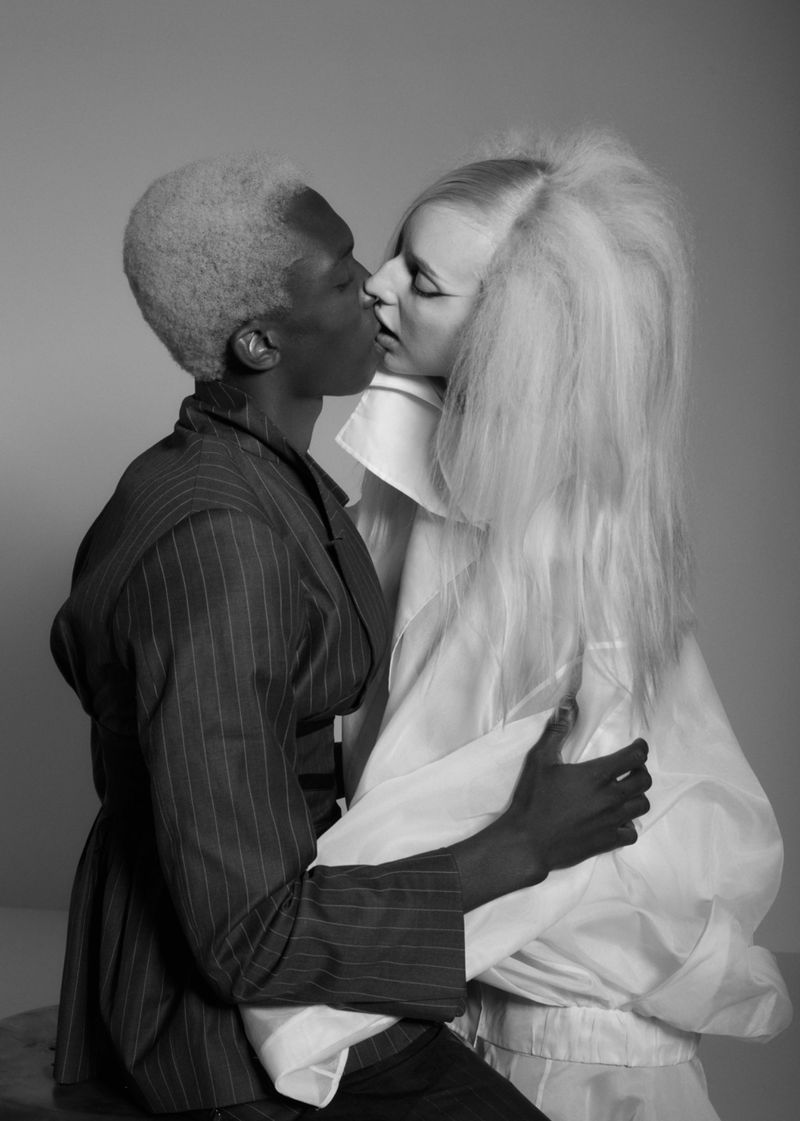 Black and white portrait of an interacial couple kissing, fashionably dressed with white and pinstripe textures in a studio setting.