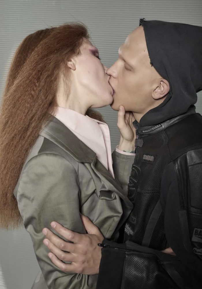 Close-up profile of a woman with crimped auburn hair in a grey blazer kissing a man wearing a black hooded moto jacket.