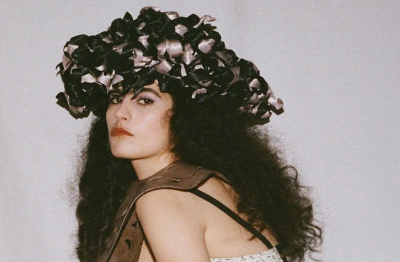 Portrait of a woman with dark curly hair and a decorative floral headpiece, wearing a brown top, against a light background.