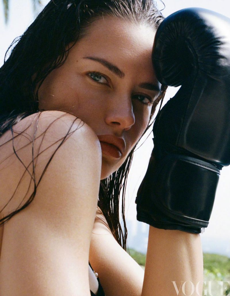 Close-up portrait of a woman with wet hair and boxing gloves, set against a blurred outdoor backdrop featuring light and airy tones.