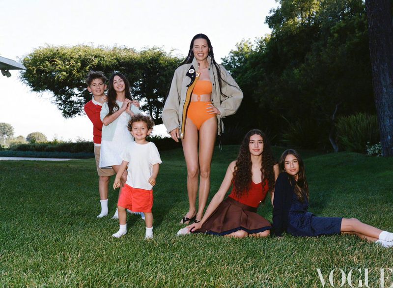 Editorial family portrait of a woman in orange swimwear with five children of mixed ages outdoors on a lawn, Vogue logo visible.