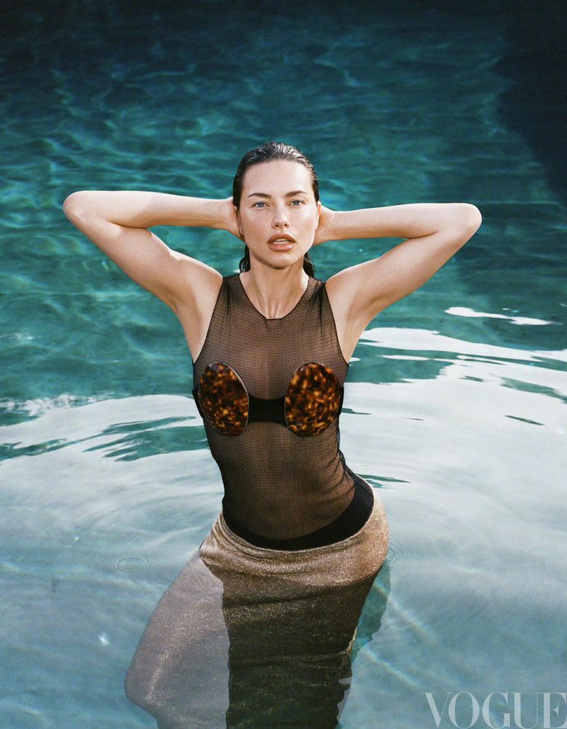 A woman in a wet black swimsuit and gold skirt stands waist-deep in a pool, with her hands behind her head in a high-angle Vogue fashion shot.