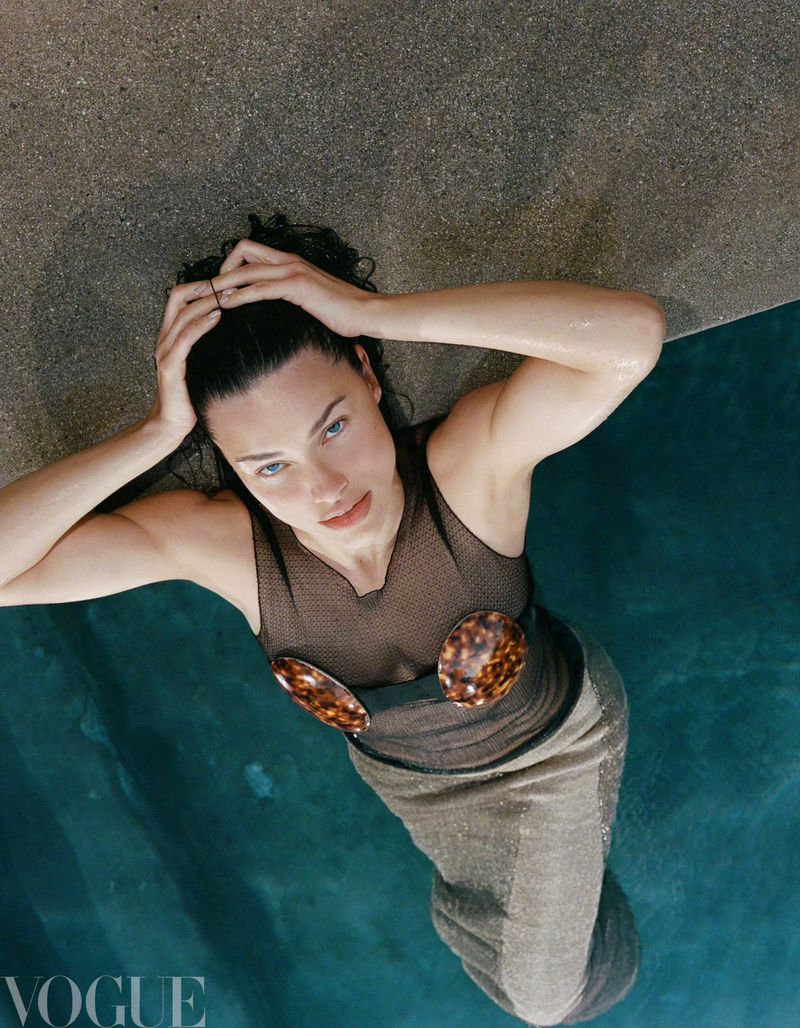 Overhead shot of Adriana Lima in water, wearing a dark top and light skirt, with hands on her head. Vogue logo visible.