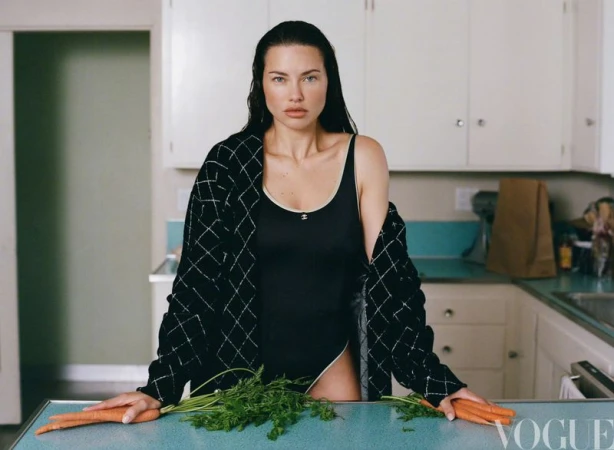 Adult woman poses indoors near carrots and a black sweater against a kitchen backdrop; the image is well-lit and professionally styled.