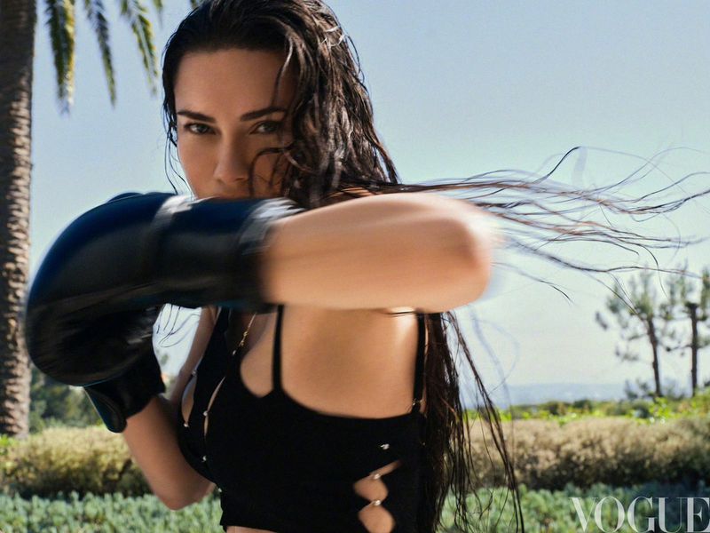 Adult woman punches towards the camera wearing black boxing gloves and an open black top, with palm trees in the background.