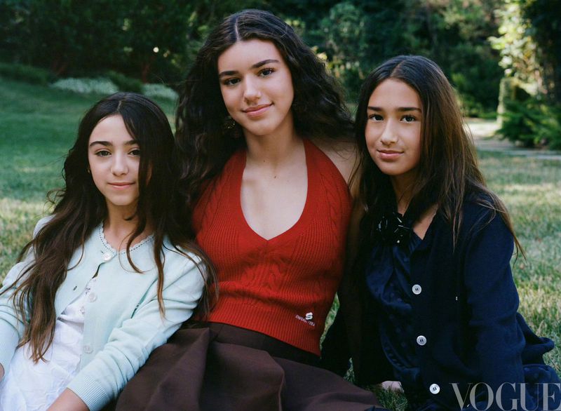 Three young women with long brown hair pose together outdoors on a grassy field; one wears a red top, another a blue jacket. Vogue logo visible.