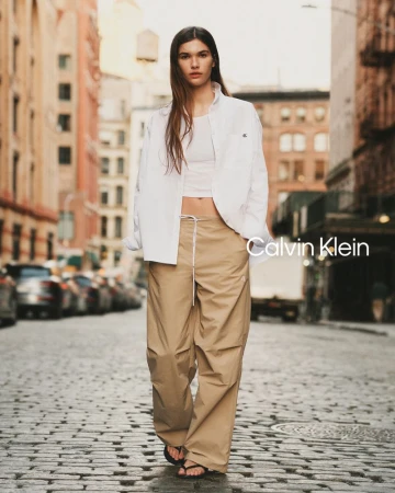 Model River Klein walking down a cobblestone city street in an open white shirt and wide-leg khaki utility pants for Calvin Klein.