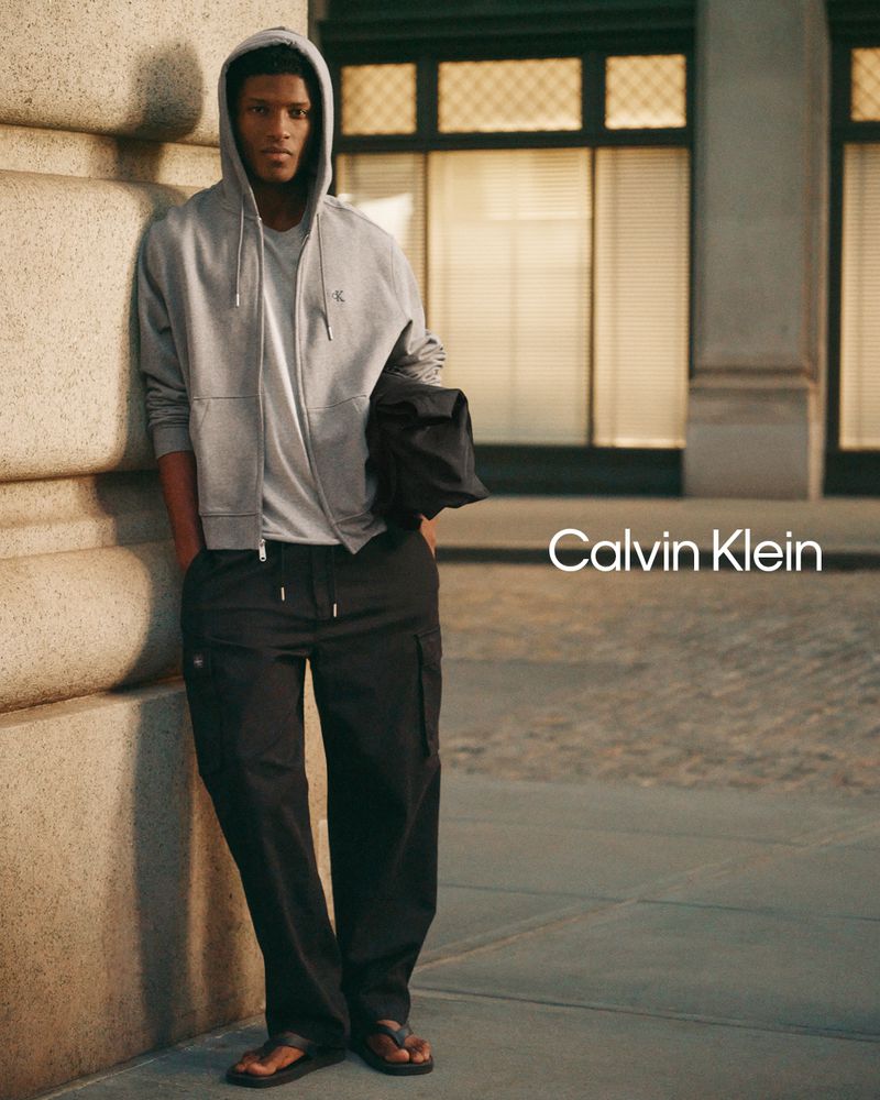 Model in grey zip hoodie and black cargo pants leaning against a stone column outdoors