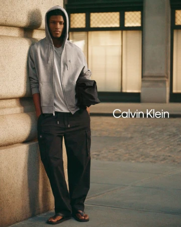 Model in grey zip hoodie and black cargo pants leaning against a stone column outdoors