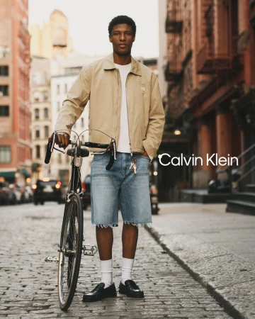 Black man standing on a cobblestone street holding a vintage bicycle, wearing a beige jacket and light denim cutoff shorts.