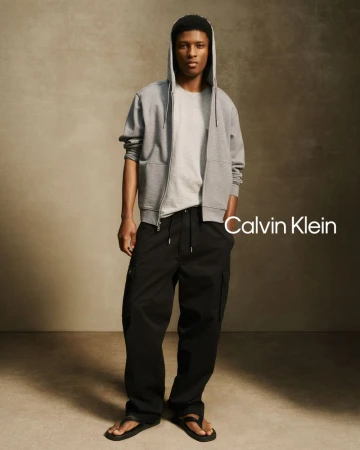 Model Aboubakar Konte wearing a grey zip hoodie and black cargo pants against a textured neutral studio backdrop for Calvin Klein.