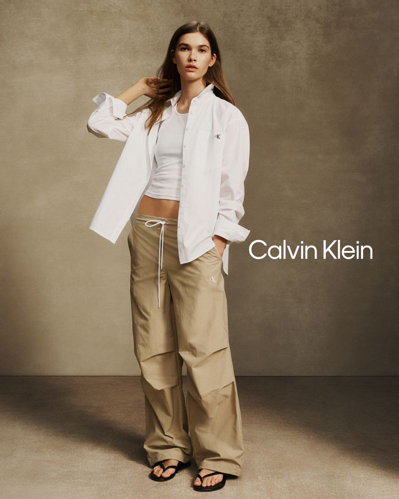 Model wearing an open white button-down shirt, white tank top, and loose beige utility trousers against a warm textured studio backdrop.