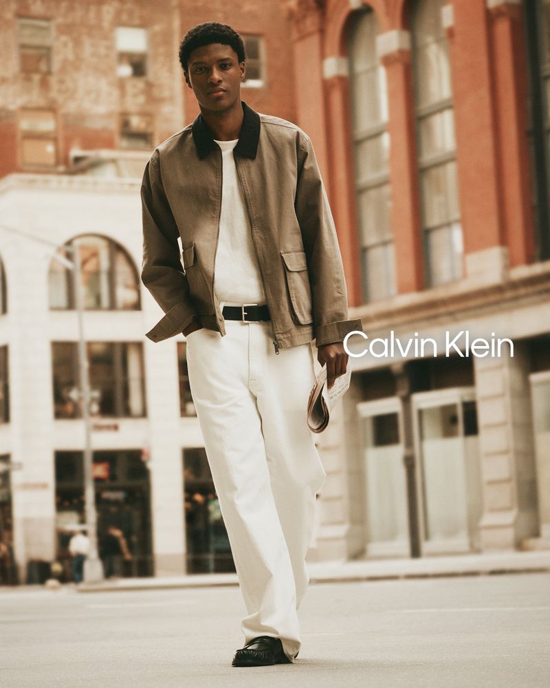 Model Aboubakar Konte walking on an urban street wearing a tan utility jacket and white trousers for Calvin Klein.