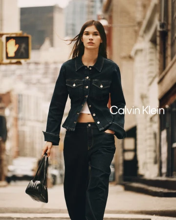 Model River Klein walking down a city street wearing a dark denim jacket, matching jeans, and holding a black shoulder bag.
