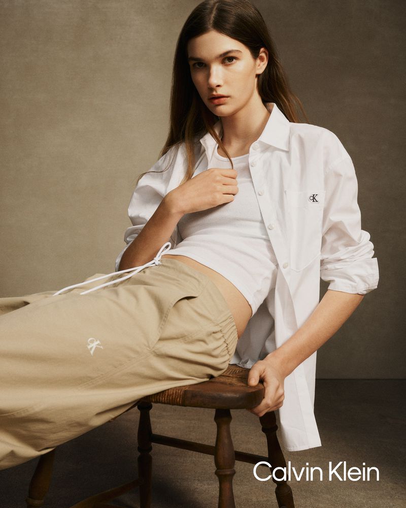 Model wearing a white shirt and khaki trousers seated on a small wooden stool against a neutral backdrop for Calvin Klein