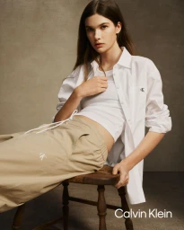 Model wearing a white shirt and khaki trousers seated on a small wooden stool against a neutral backdrop for Calvin Klein