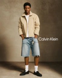 Black male model wearing a beige utility jacket and denim cutoff shorts against a warm neutral studio backdrop.