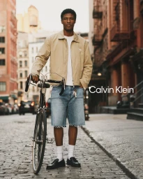 Black man standing on a cobblestone street holding a vintage bicycle, wearing a beige jacket and light denim cutoff shorts.