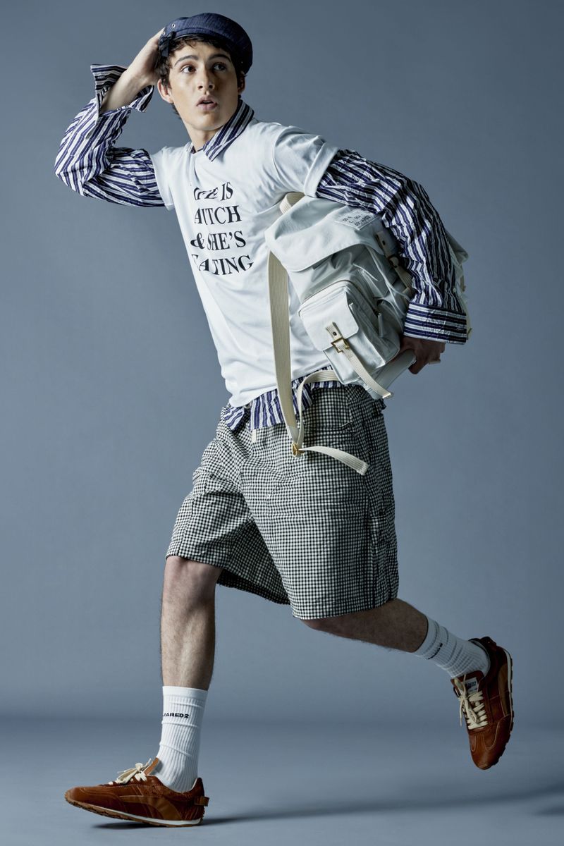 A young adult male with fair skin in shorts, a striped shirt, and a T-shirt is running with a backpack against a studio backdrop.