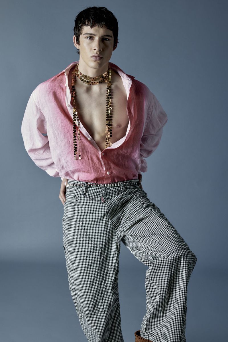 Young adult male model in a pink ombre shirt, checkered pants, and golden necklace poses in a fashion forward studio shot.
