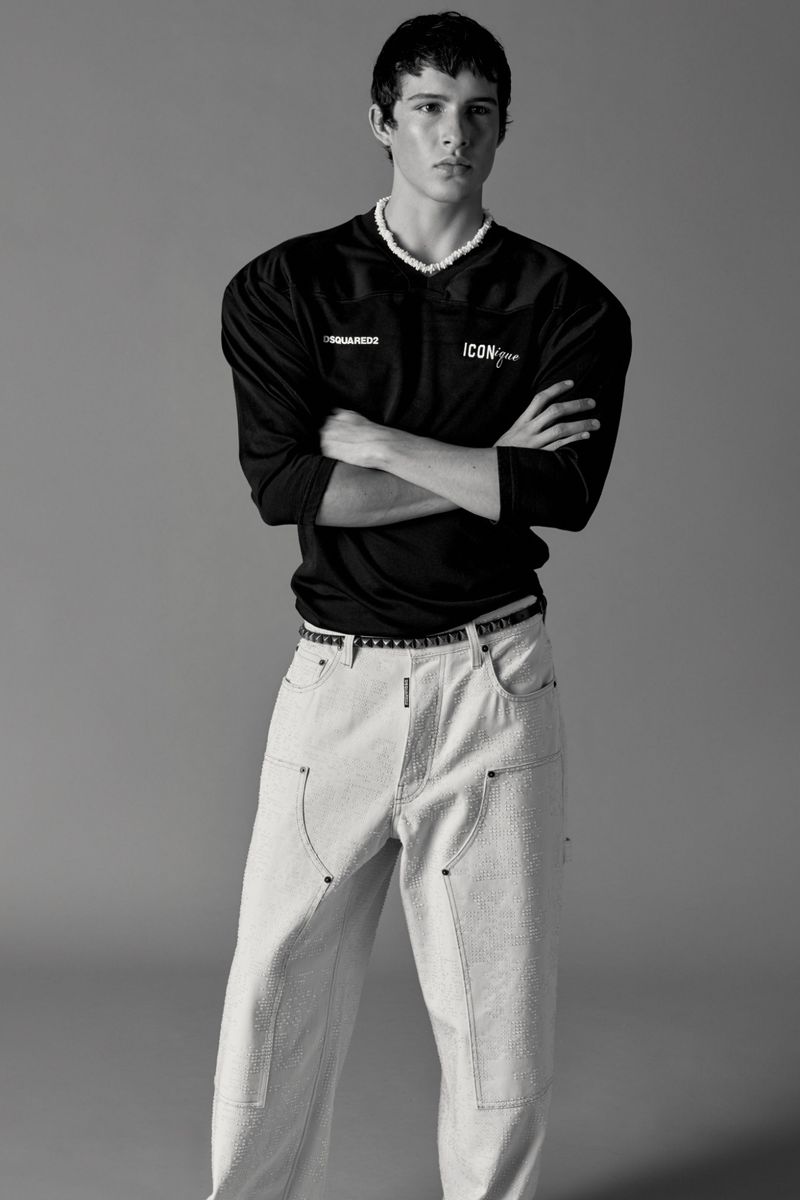 Monochrome fashion portrait of a young adult male wearing a black long-sleeved shirt and light-colored jeans posing indoors.