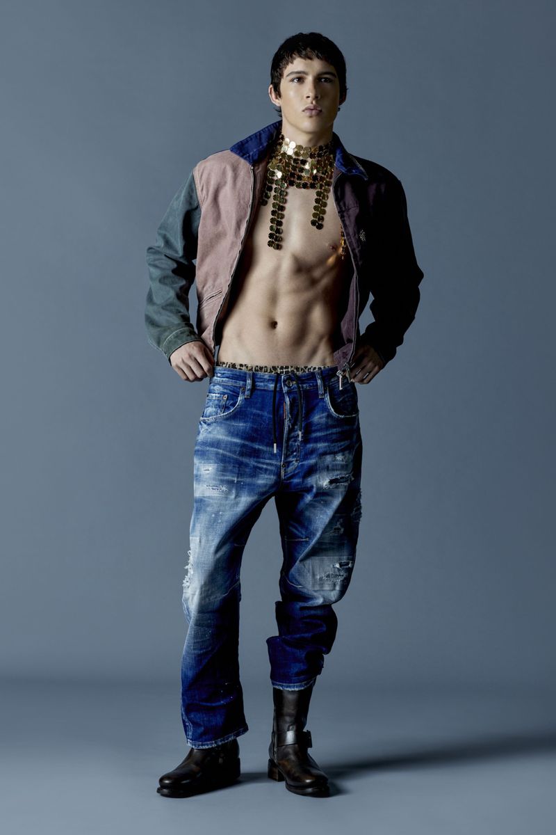 Full shot of a fit adult male in distressed jeans, boots and jacket with a metallic bib posing in a studio with a grey backdrop.