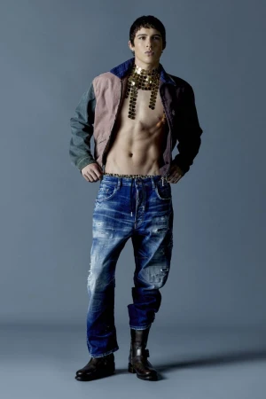 Full shot of a fit adult male in distressed jeans, boots and jacket with a metallic bib posing in a studio with a grey backdrop.