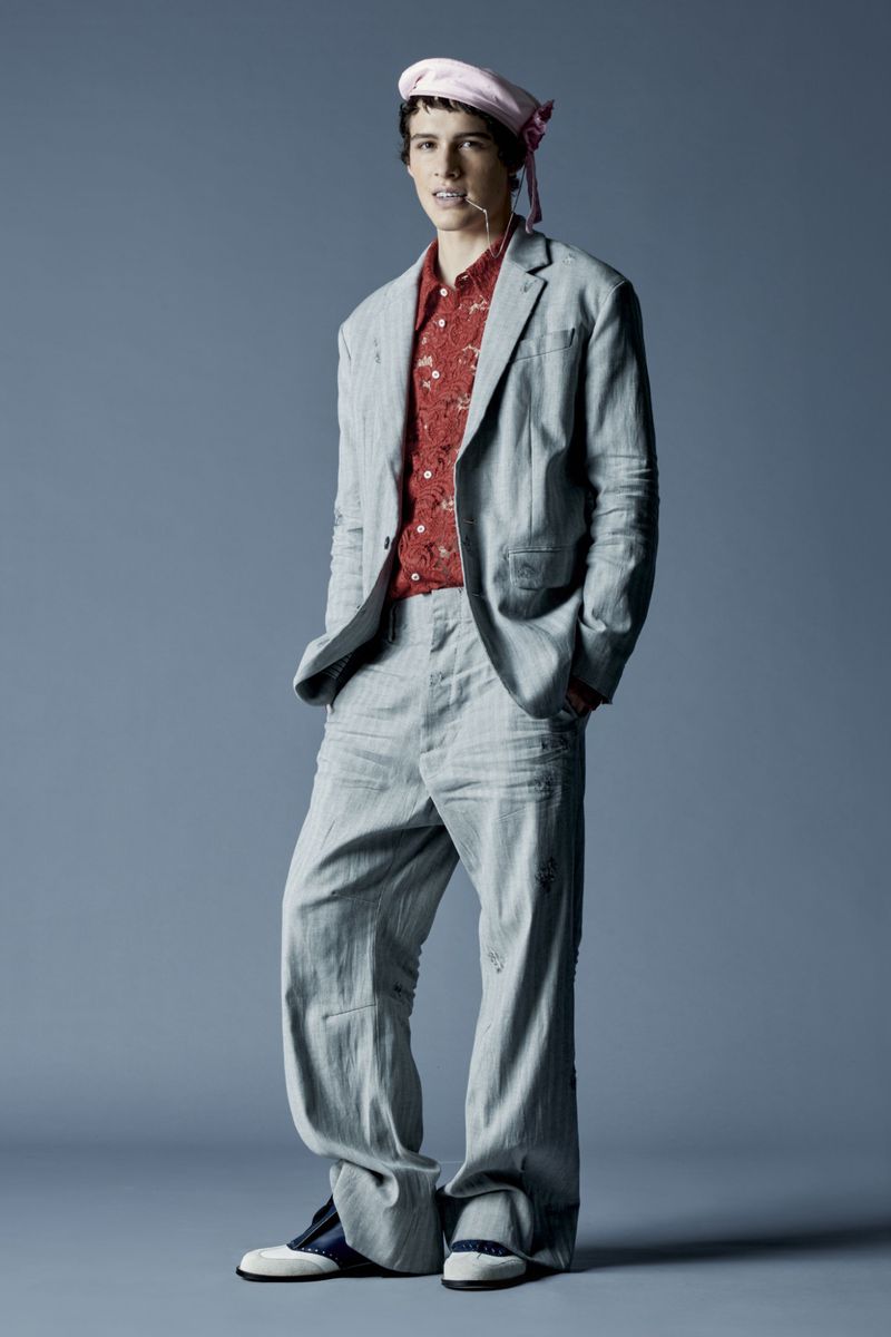 Full shot of a young man in an oversized gray suit and pink hat standing in a photo studio with a gray background.