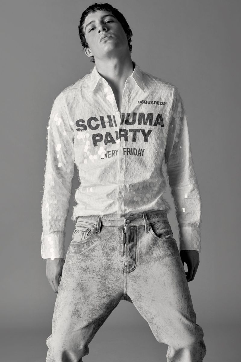 Monochrome portrait of a young adult man in a sequined button-down shirt and jeans, posing against a plain grey backdrop in a studio setting.