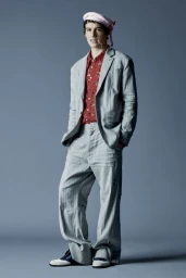 Model wearing a grey suit jacket, loose trousers, patterned red shirt, and a pink hat against a grey studio backdrop.