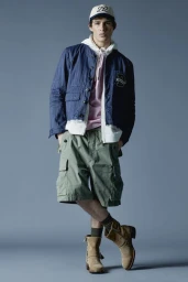 Male model wearing a blue jacket, white hoodie, pink shirt, khaki shorts, and tan boots against a grey studio backdrop