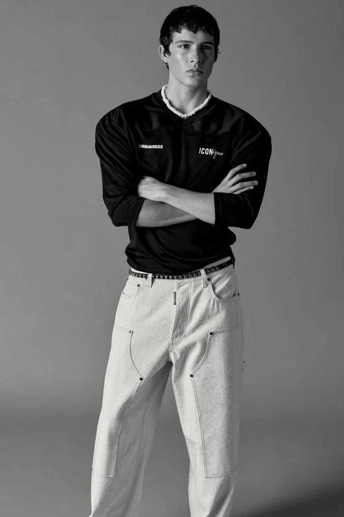 Male model Brennan Aldred posing with arms crossed in a black Dsquared2 jersey and light wide-leg jeans against a grey backdrop.