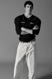 Male model Brennan Aldred posing with arms crossed in a black Dsquared2 jersey and light wide-leg jeans against a grey backdrop.