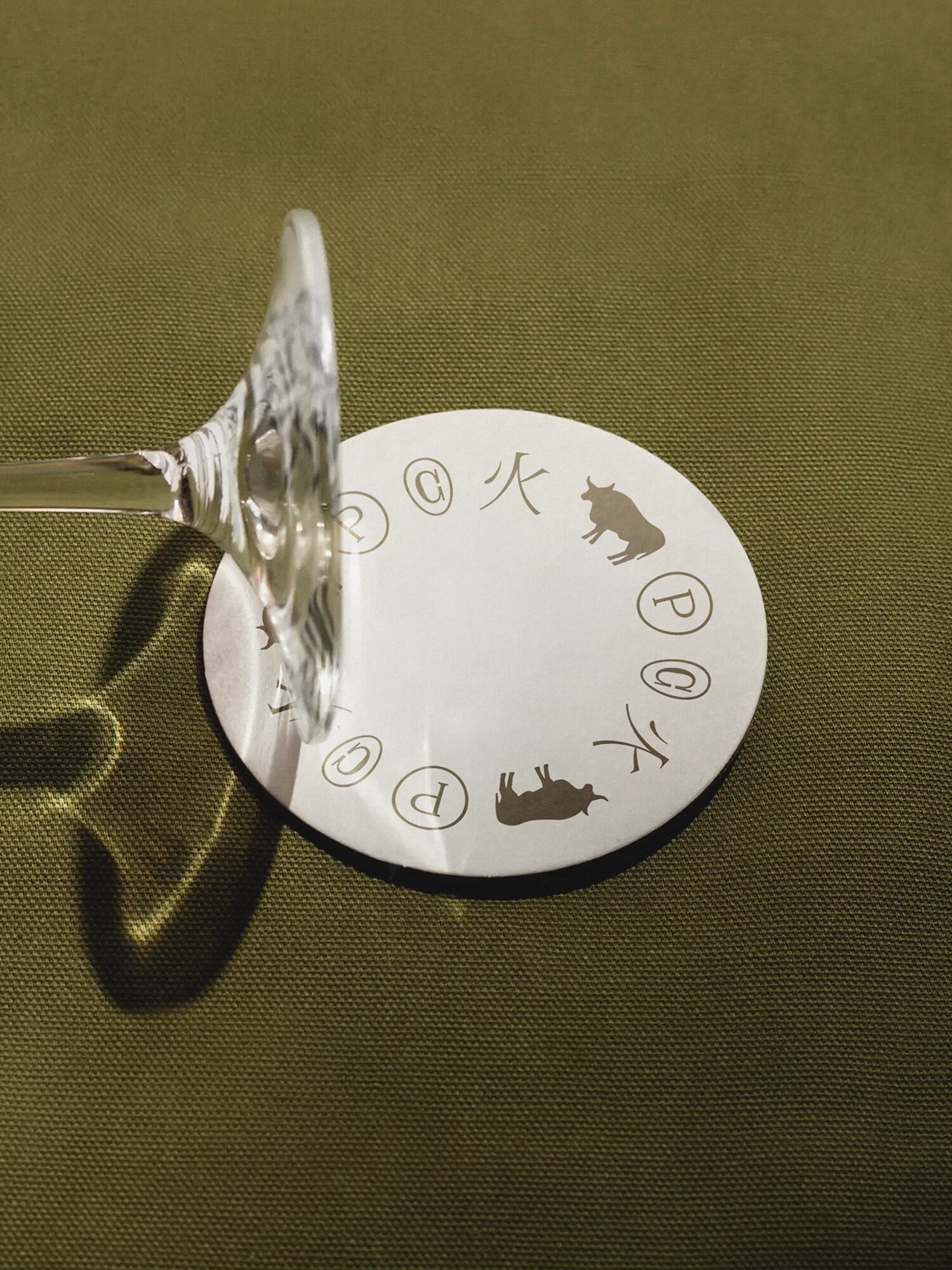 A white circular coaster with printed icons under the base of a clear wine glass on olive green textured fabric.