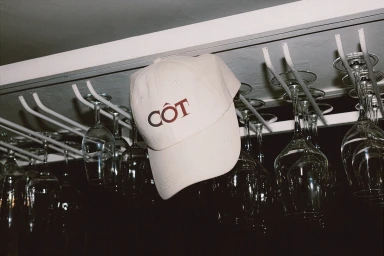 Cream baseball cap with burgundy embroidered Côt logo hanging from a metal overhead wine glass rack in a bar.