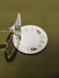 A white circular coaster with printed icons under the base of a clear wine glass on olive green textured fabric.