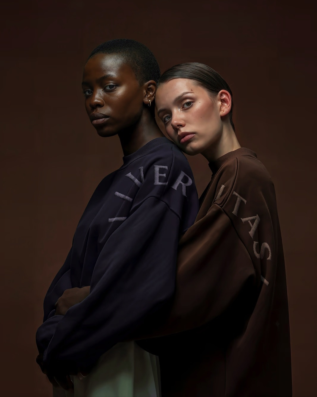 Two female models in navy and brown oversized Veritas sweatshirts against a dark brown studio background.
