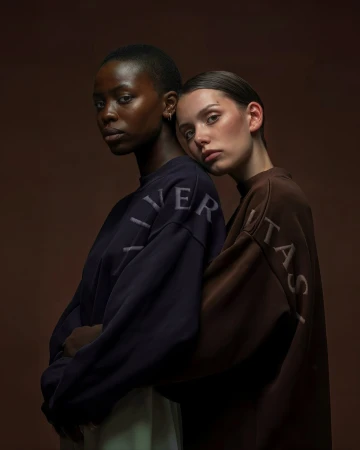 Two female models in navy and brown oversized Veritas sweatshirts against a dark brown studio background.