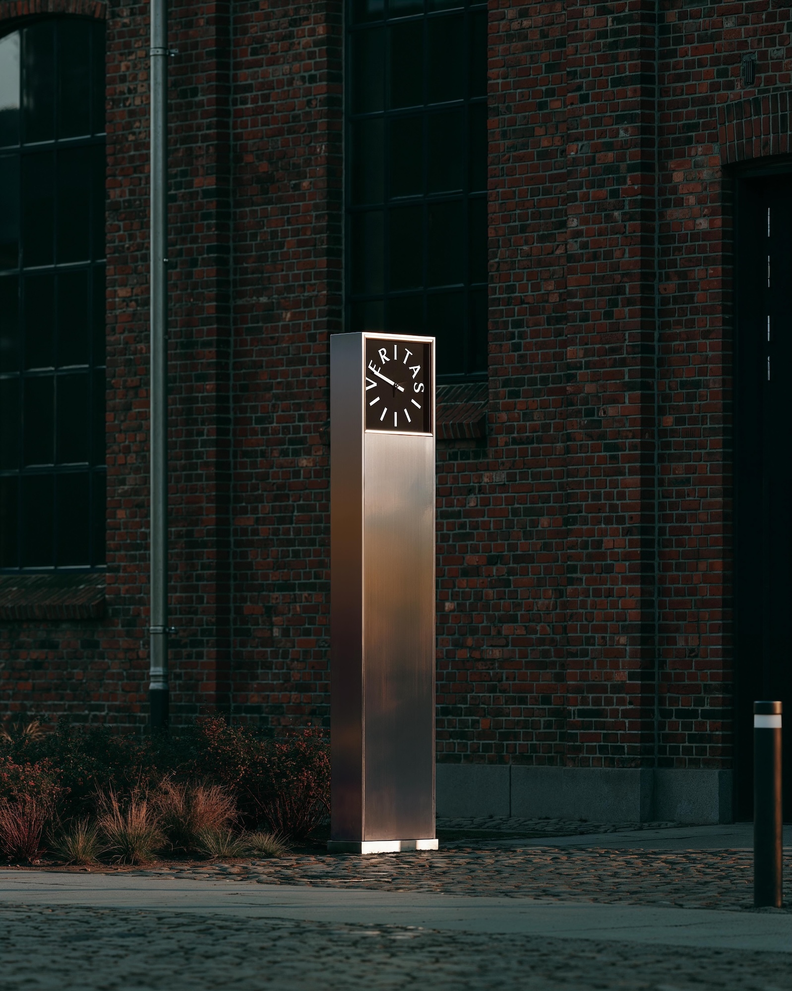 Brushed metal totem clock with illuminated Veritas face standing outside a red brick building on stone pavement.