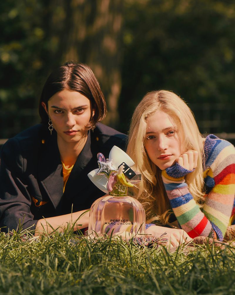 Two models lying on grass outdoors next to a Marc Jacobs Perfect perfume bottle in a campaign shot by Leon Mark.