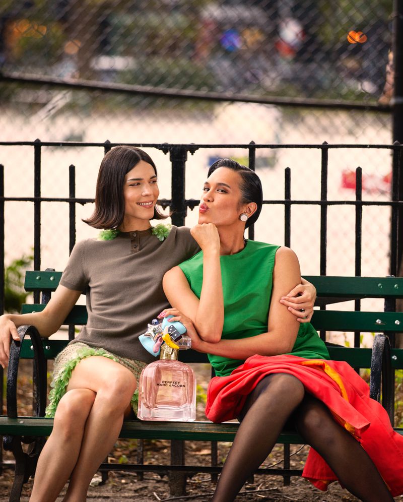 Two models sitting closely together on a green park bench beside an oversized bottle of Marc Jacobs Perfect perfume.