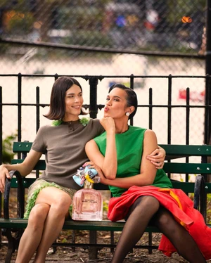 Two models sitting closely together on a green park bench beside an oversized bottle of Marc Jacobs Perfect perfume.