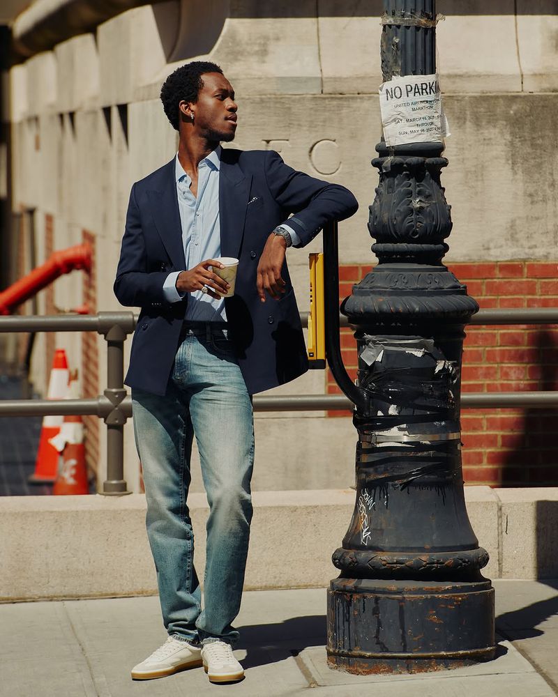 A stylish black man in a navy blazer, light blue shirt, and jeans, stands against a city lamppost, holding a coffee cup, during daytime.