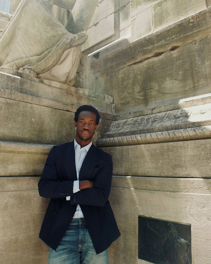 Portrait of African American man in blue blazer and jeans, arms crossed, leaning against a stone wall outdoors in daylight.