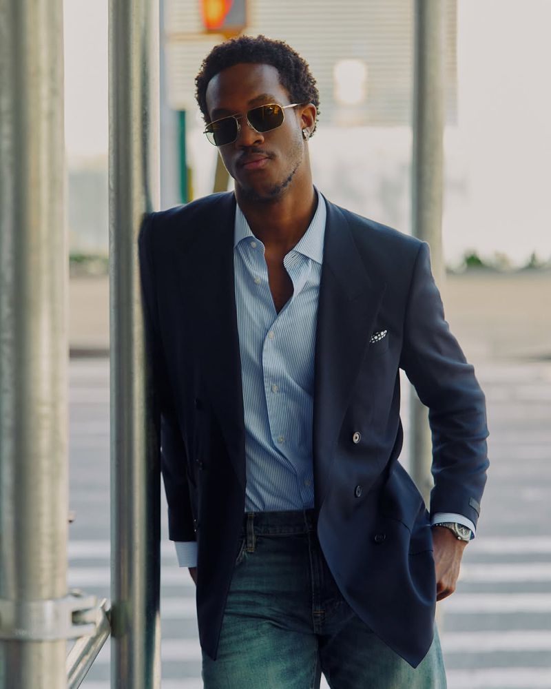 Stylish African-American man in a navy blazer and sunglasses leans against a city pole, looking confident against an urban backdrop.