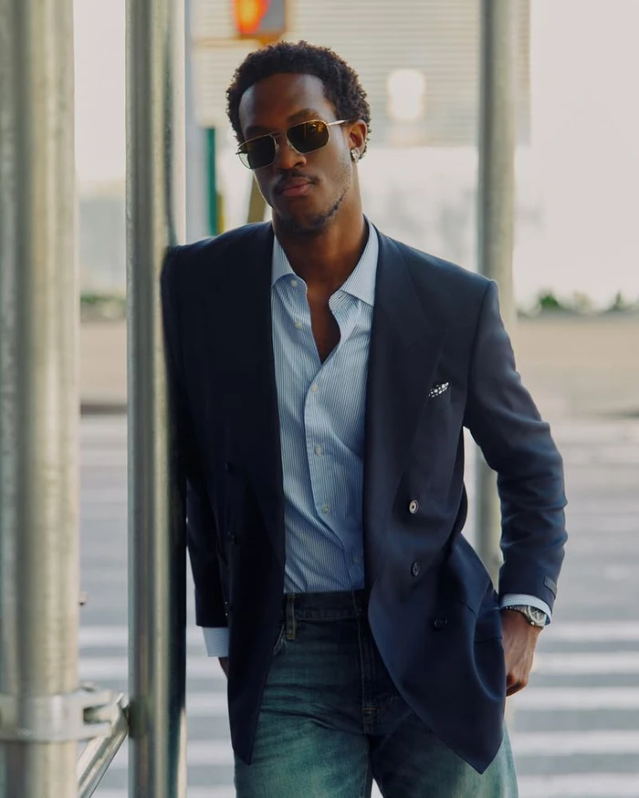 Wisdom Kaye wearing sunglasses and a navy double-breasted blazer with a blue shirt while leaning against a metal post on a city street.