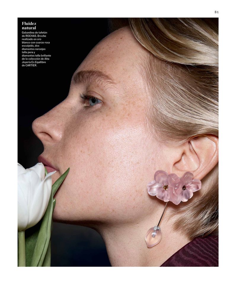 Close-up studio shot of a blond woman with freckles wearing a pink flower earring, holding a tulip near her lips in a beauty editorial.